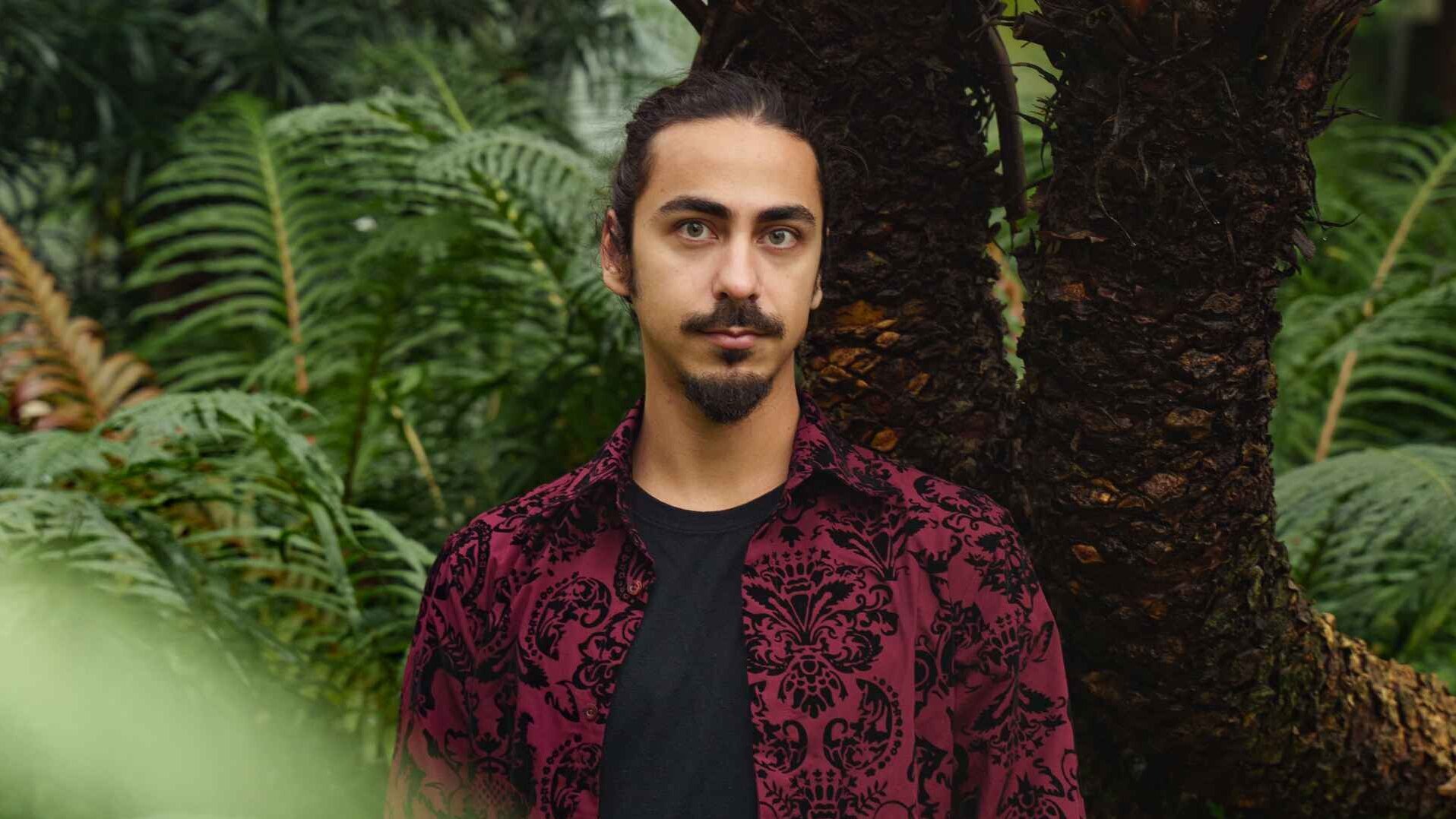 Elia Hussain steht in einem dunkelroten Jackett einem Wald von Palmen