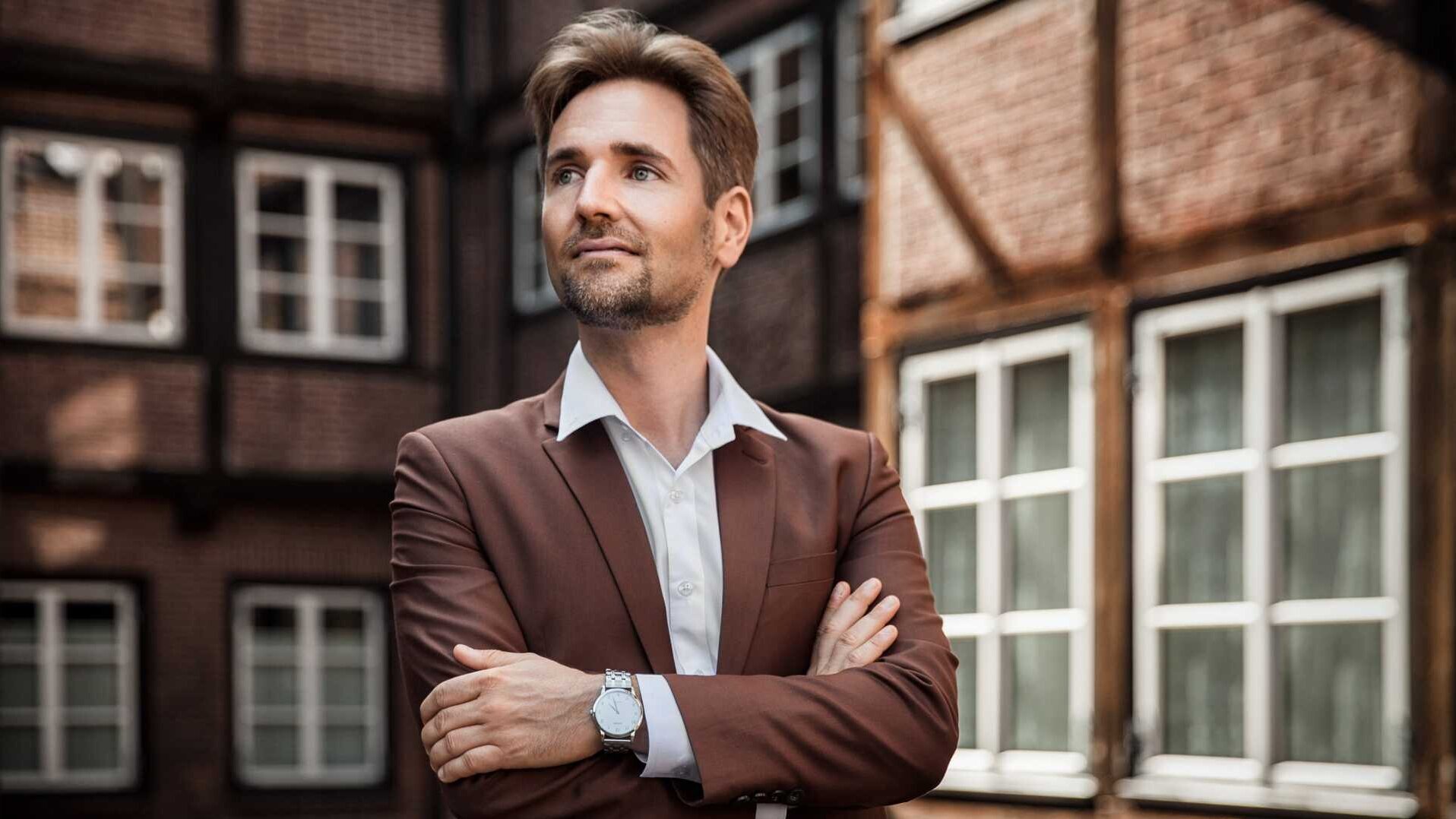 Laurens Patzlaff steht in einem braunen Anzug vor einem Fachwerkhaus